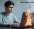 A man sitting at a desk using a laptop that is overheating, with smoke and flames rising from the keyboard, while he looks frustrated and upset.