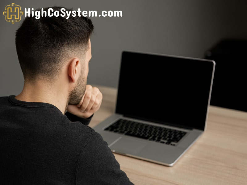 A person sits at a desk facing a laptop with a black screen, hand under chin, thinking about the issue